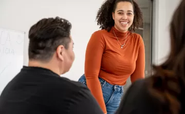 Woman in orange long sleeve shirt smiles as she stands in front of small group in a classroom setting