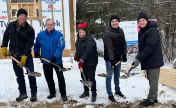 Five people hold shovels with dirt in front of the frame of a building