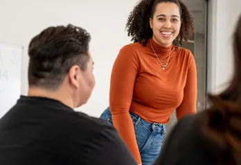 Woman in orange long sleeve shirt smiles as she stands in front of small group in a classroom setting