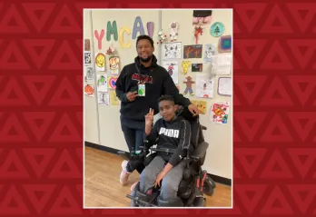 A boy in a wheelchair holds up the peace sign, a young man stands behind him also doing the peace sign 
