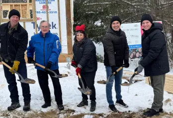 Five people hold shovels with dirt in front of the frame of a building