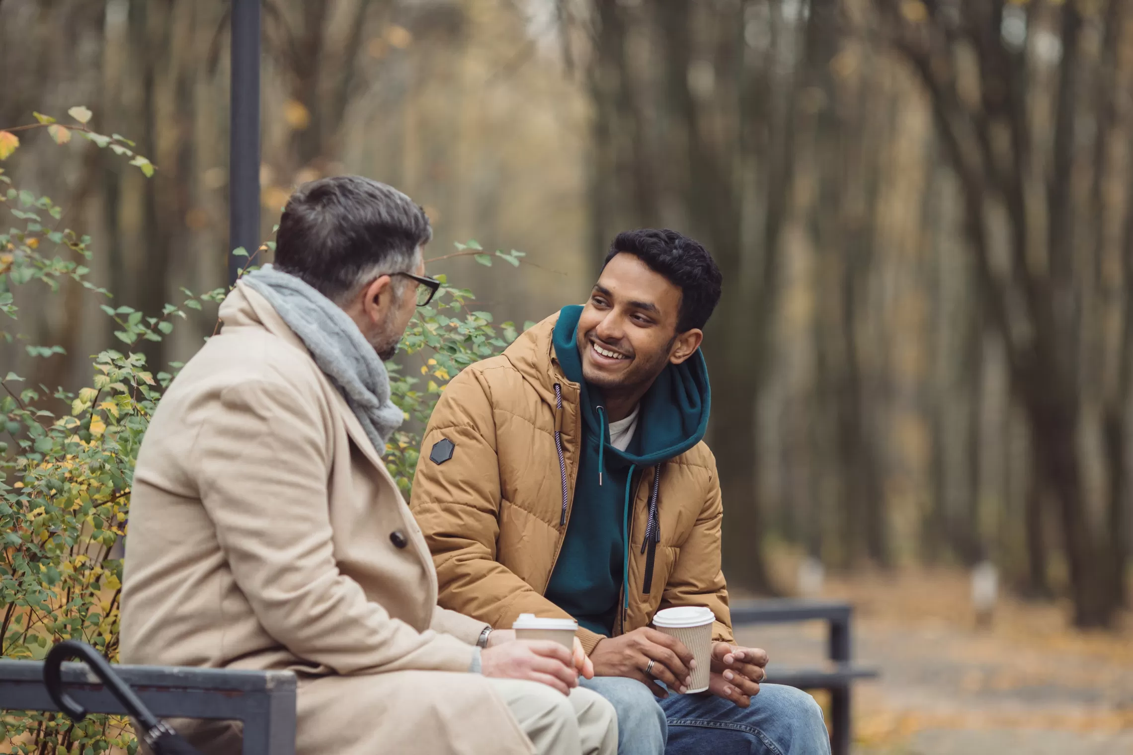 Two men are sitting on a bench in the fall. They are smiling, drinking coffee and having a discussion. 