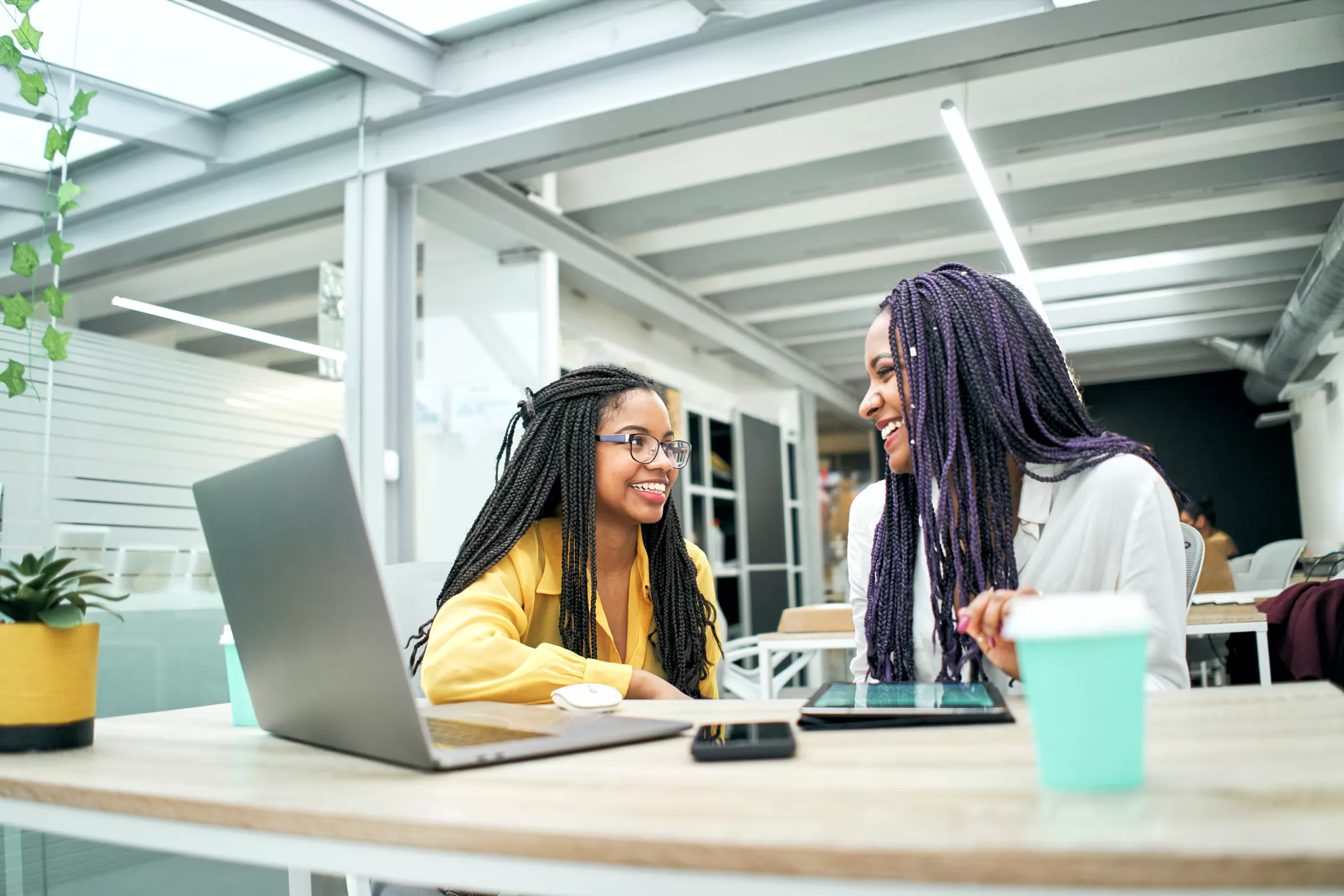 Two women are smiling talking in front of laptop. Both have braids, one is younger than the other. There is a coffee cup on the table.