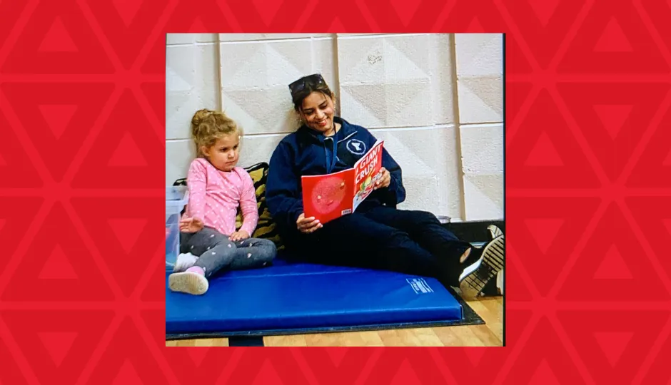 A early childhoos educator sits on a blue mat on a gymnasium floor reading a red book to a little girl in a pink shirt.