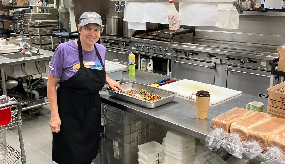 strong, elderly woman with short grey hair standing in a commercial kitchen prepping vegetables