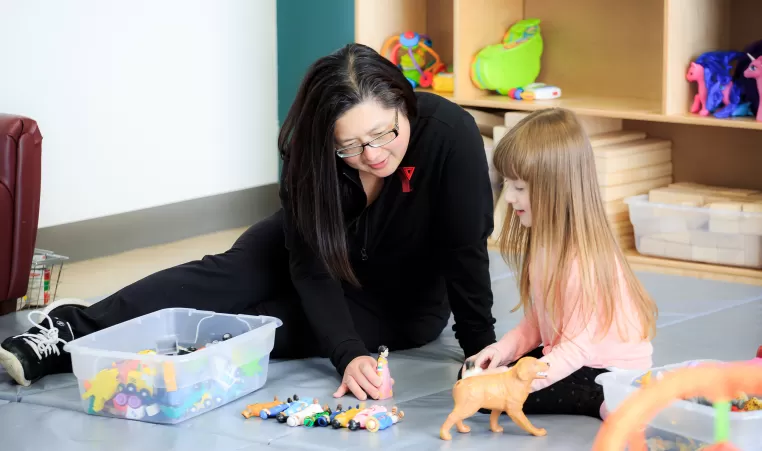 YMCA Child Care Educator and female child playing with animal figures on floor mat