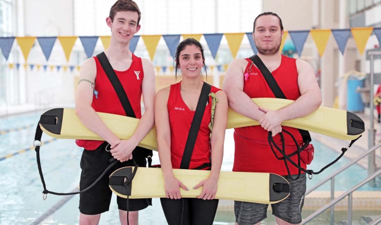 An image of two males and one female YMCA lifeguards smiling as they hold on to their safety equipment in front of a pool.