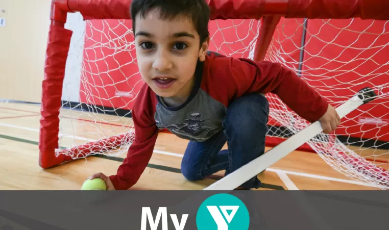 A young boy looking at the camera while down on one knee in a hockey net. In his right hand a hockey stick and his left a tennis ball.