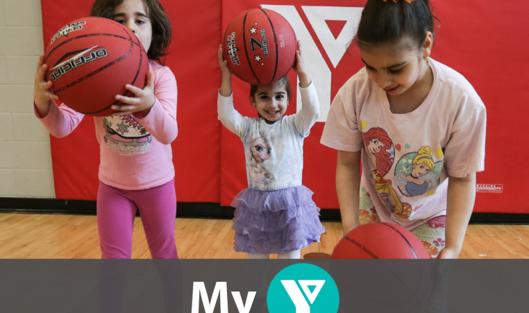 Three young girls play with basketballs inside the YMCA gym.