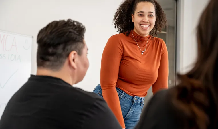 Woman in orange long sleeve shirt smiles as she stands in front of small group in a classroom setting