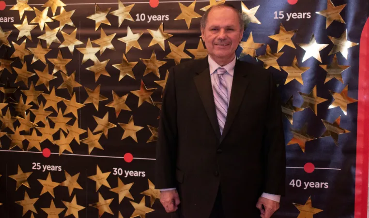 Older gentleman in suit stands in front of board with gold stars