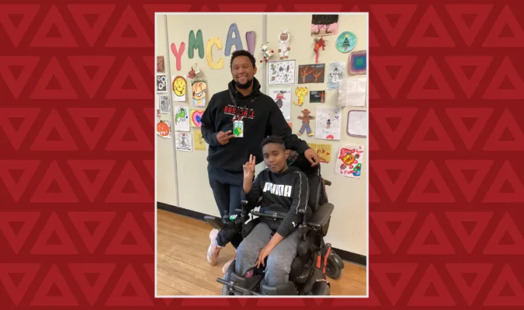 A boy in a wheelchair holds up the peace sign, a young man stands behind him also doing the peace sign 