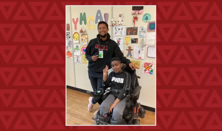 A boy in a wheelchair holds up the peace sign, a young man stands behind him also doing the peace sign