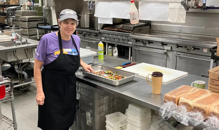 strong, elderly woman with short grey hair standing in a commercial kitchen prepping vegetables