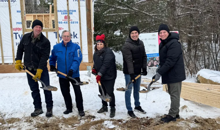 4 men and 1 women hold shovels with dirt in front of the frame of a building