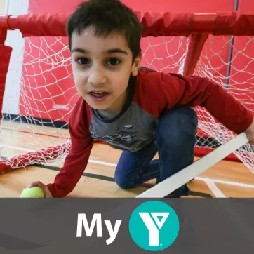 A young boy looking at the camera while down on one knee in a hockey net. In his right hand a hockey stick and his left a tennis ball.
