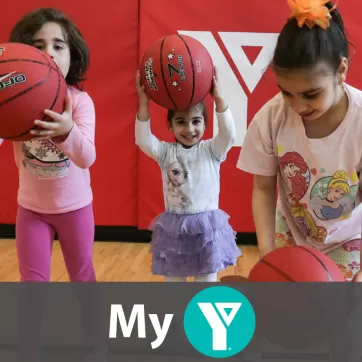 Three young girls play with basketballs inside the YMCA gym.