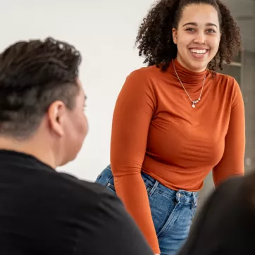 Woman in orange long sleeve shirt smiles as she stands in front of small group in a classroom setting