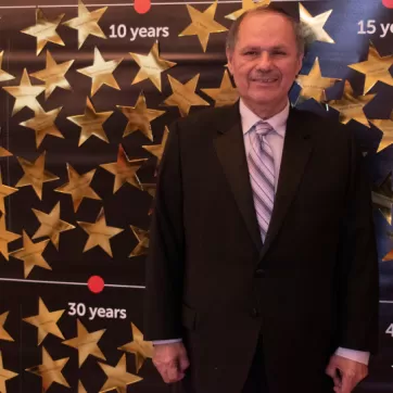 Older gentleman in suit stands in front of board with gold stars