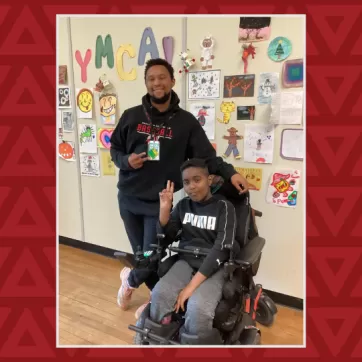 A boy in a wheelchair holds up the peace sign, a young man stands behind him also doing the peace sign 