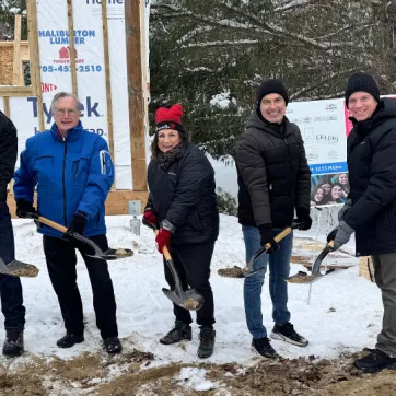 4 men and 1 women hold shovels with dirt in front of the frame of a building