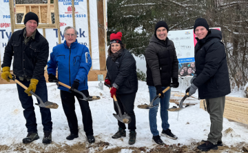 Five people hold shovels with dirt in front of the frame of a building